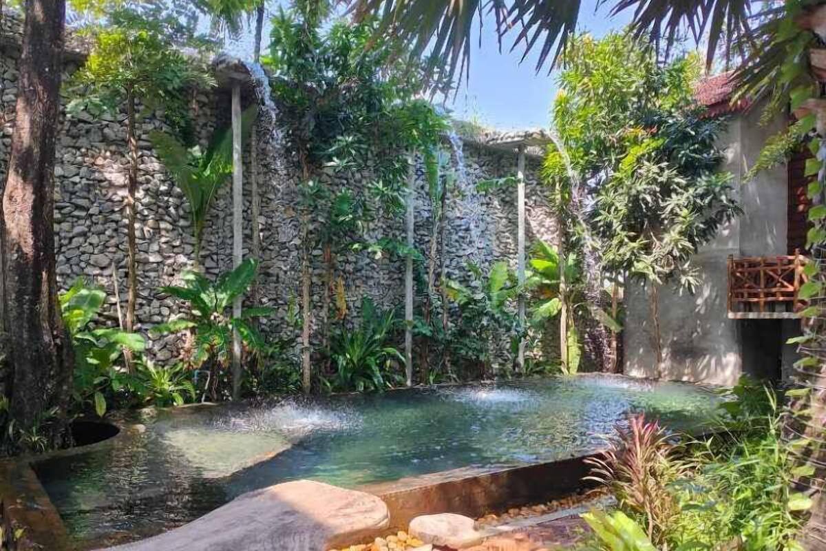 A serene outdoor pool area with a stone wall, lush greenery, and a small waterfall feature.