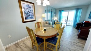 A dining area with a round table and chairs, a framed painting on the wall, and a view of the balcony through the window. - Harbor House B6 2 Bedroom Condo by RedAwning (Gulf Shores)