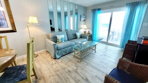 A living room with a sofa, a glass coffee table, and a chair. There is a window with curtains, a lamp, and a framed picture on the wall. - Harbor House B6 2 Bedroom Condo by RedAwning (Gulf Shores)
