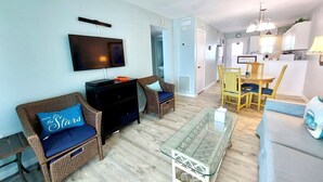 A living room with a flat-screen TV, wicker furniture, a glass coffee table, and a kitchen area in the background. - Harbor House B6 2 Bedroom Condo by RedAwning (Gulf Shores)