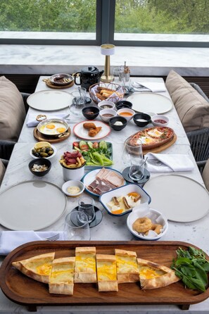 Ein Frühstückstisch ist reichhaltig gedeckt mit Speisen, darunter Eier, Fleisch und Gemüse, in einem Raum mit großem Fenster und bequemen Sitzmöglichkeiten.