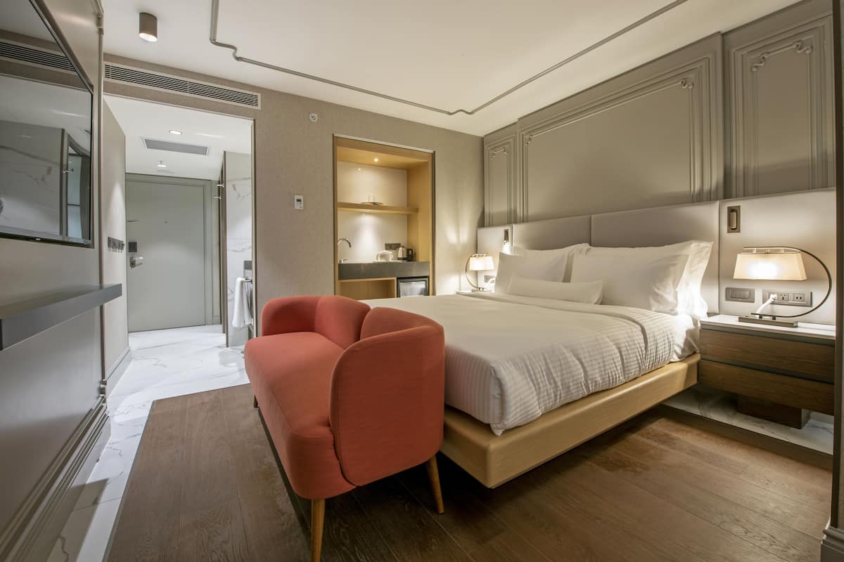 A modern hotel room with a large bed, a sitting area with a red chair, a bedside table with a lamp, and a bathroom visible through an open door.