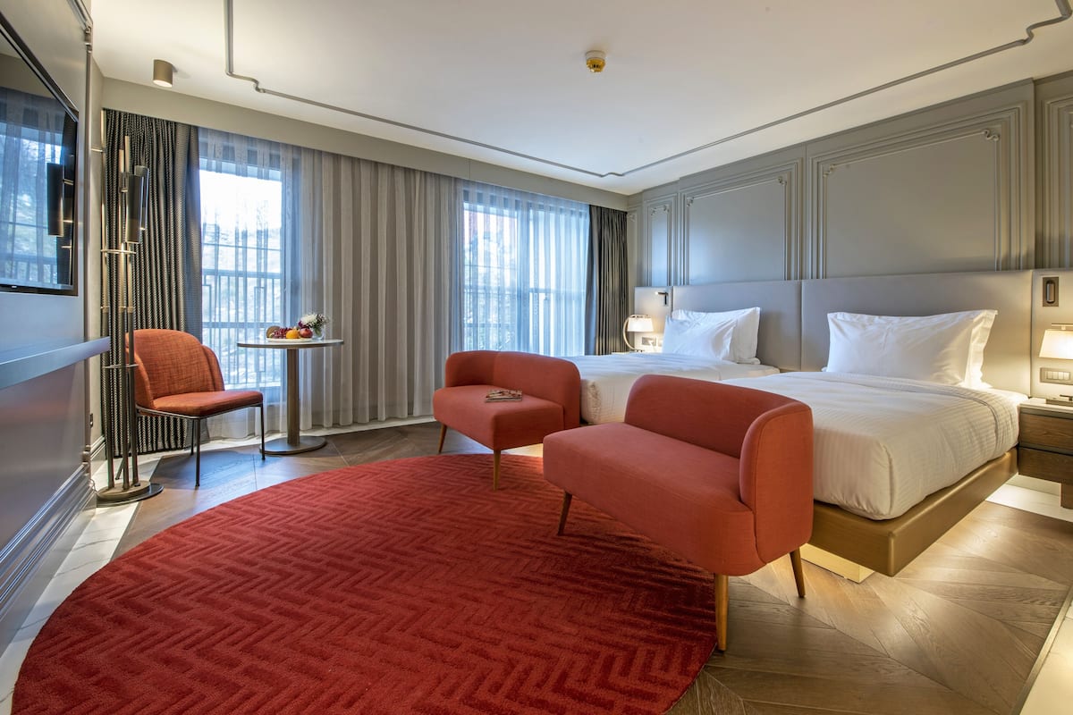 A modern hotel room with a large bed, two armchairs, a small table with fruit, and a red carpet.
