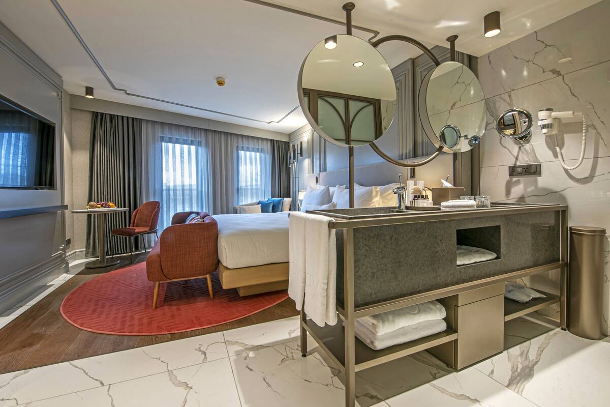 A modern hotel room with a bed, a seating area with a chair, a vanity with a mirror, and a bathroom with a toilet and sink.
