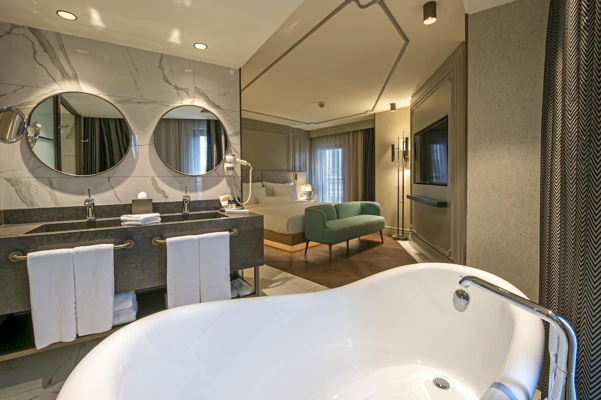 A modern hotel bathroom with a large bathtub, dual sinks, and a green armchair.