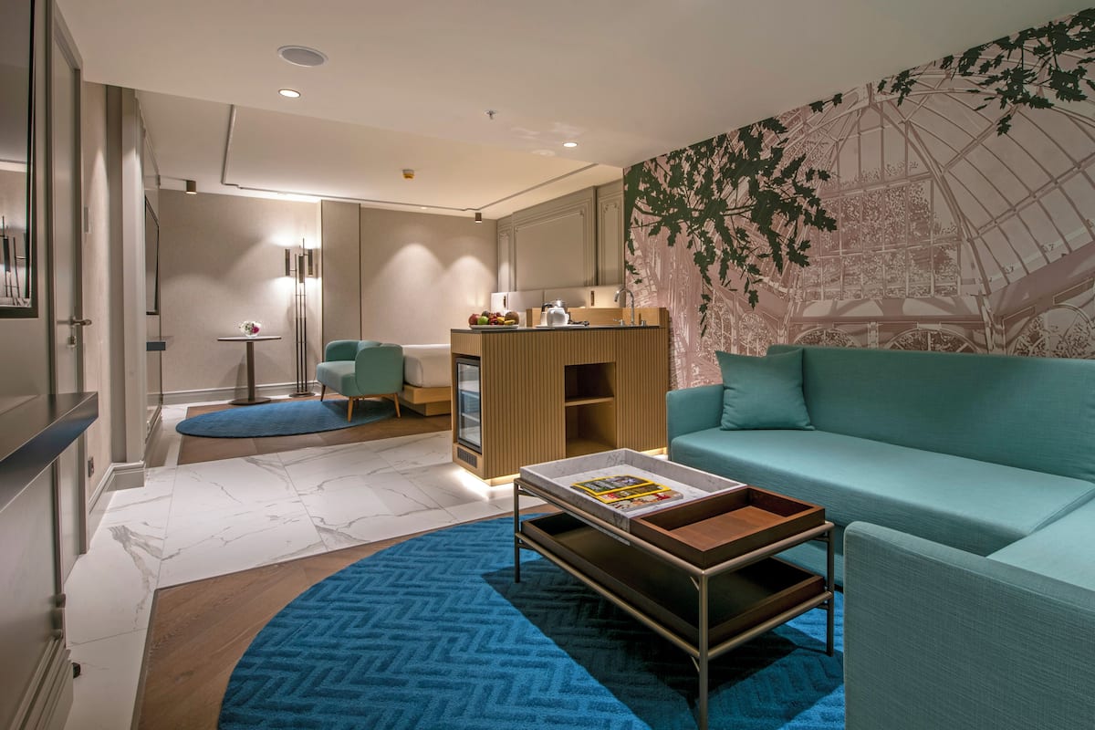 A modern living room with a teal sofa, a coffee table, a small kitchenette, and a large wall mural.