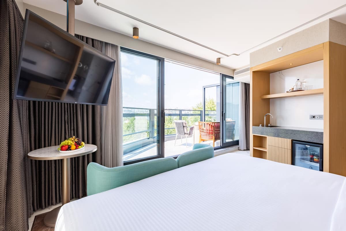 A modern hotel room with a large bed, a TV mounted on the wall, a small table with fruit, and a balcony with a chair.