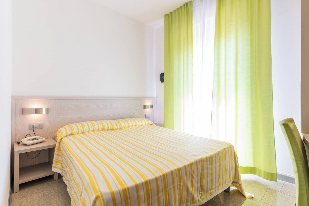 Una habitación de hotel con cama, mesita de noche, lámpara y cortinas.