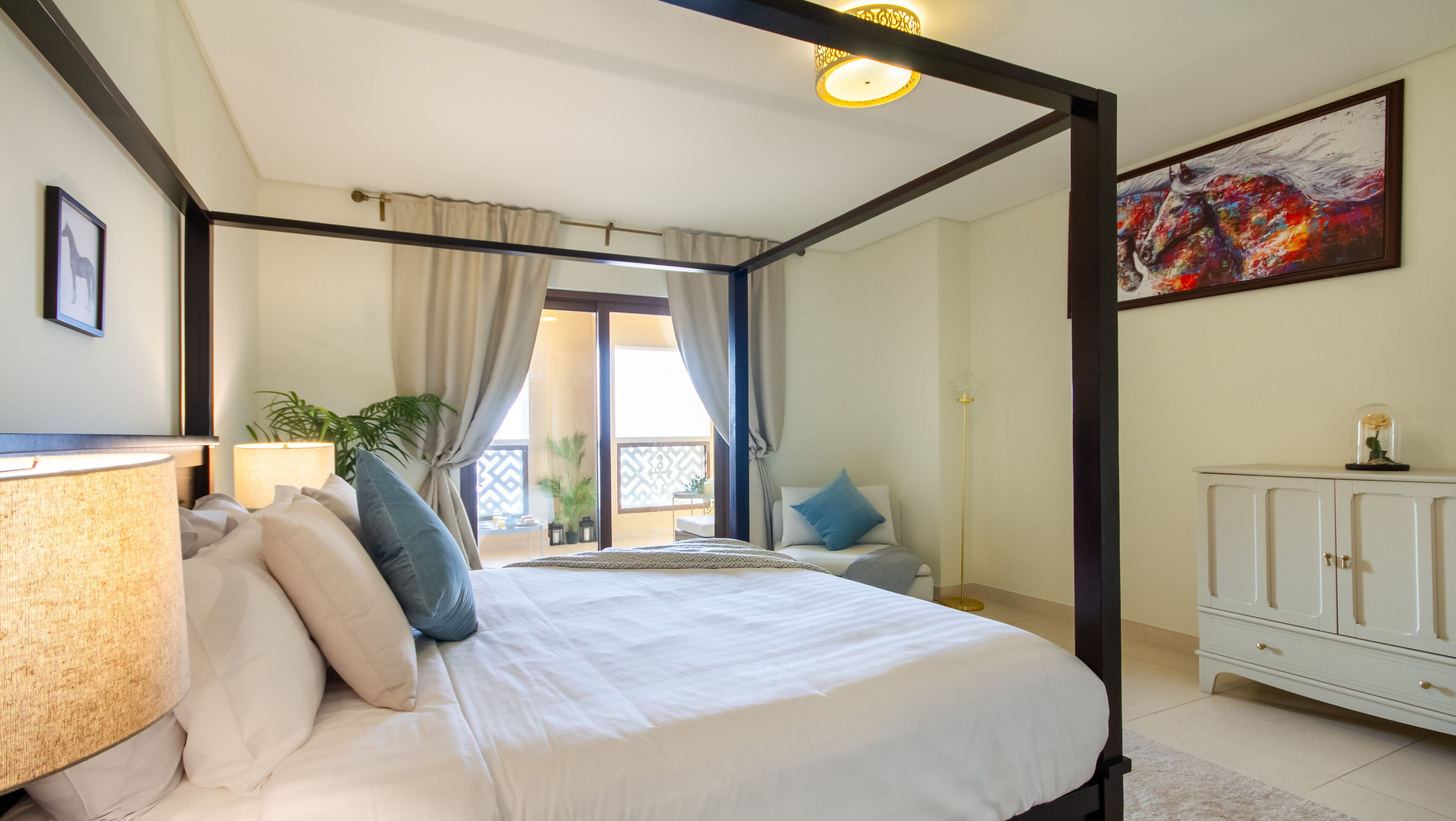 Una cama con dosel con cubrecama blanco, una mesita de noche con lámpara, vista a un balcón y un armario blanco con un adorno encima.