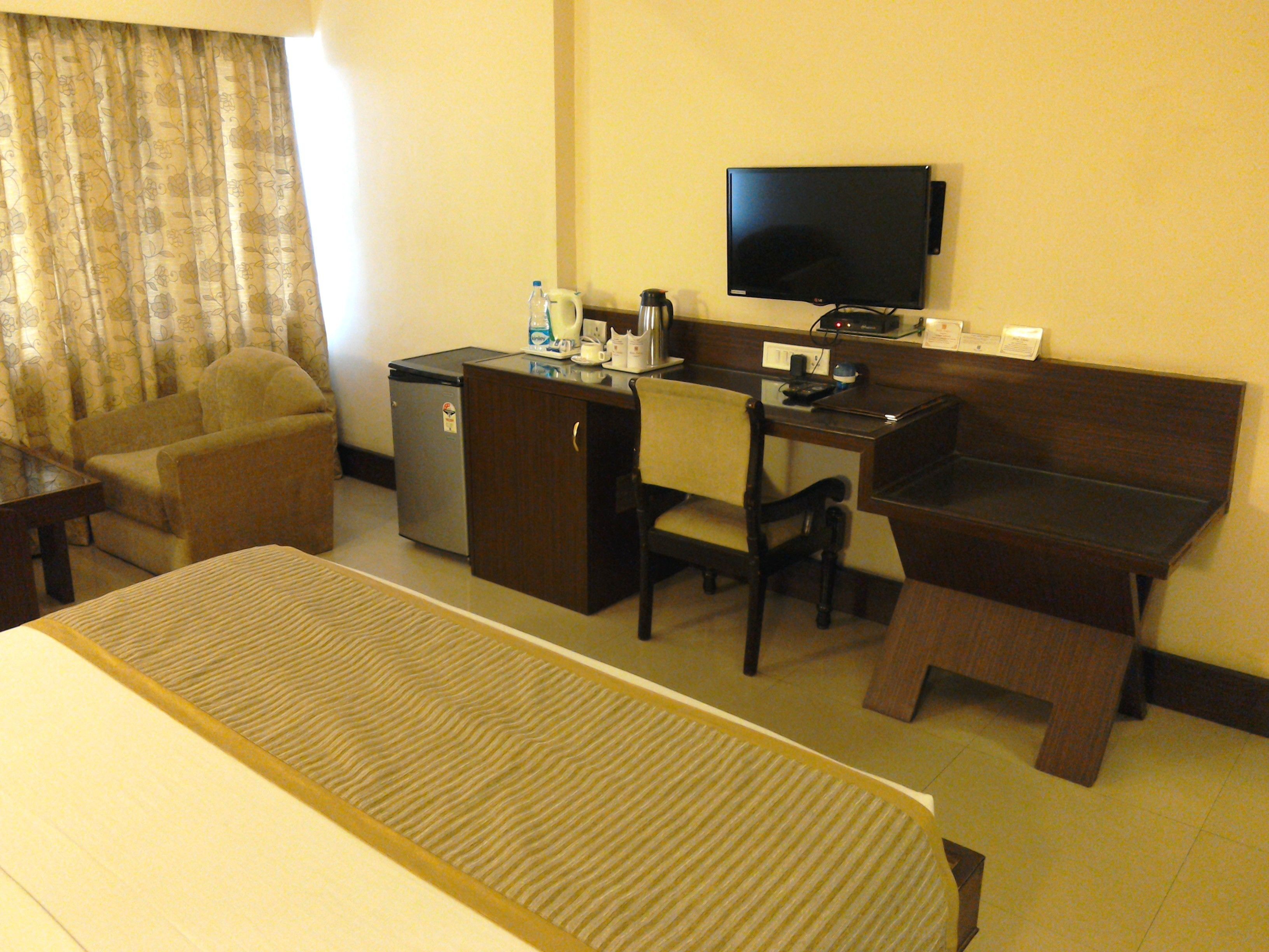 Habitación de hotel con cama, escritorio, silla, televisor, refrigerador y ventana con cortinas.