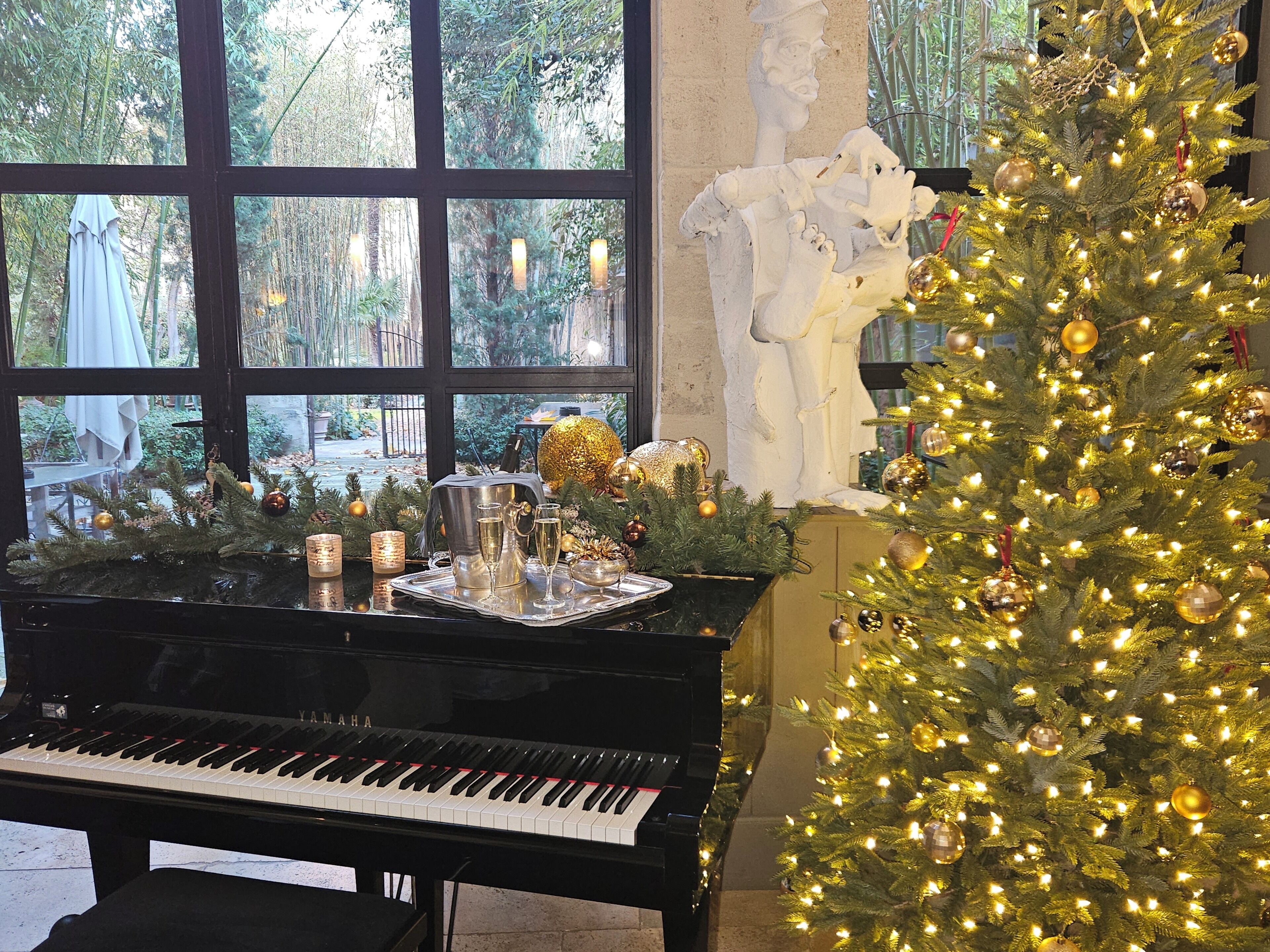 A piano, a Christmas tree, and a statue in a room with large windows.