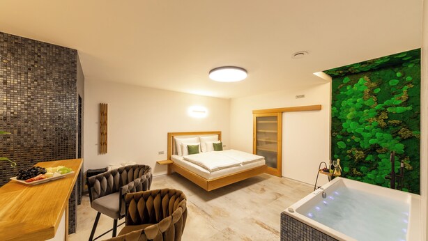 Un dormitorio moderno con una cama, un escritorio de madera con una bandeja de frutas, dos sillones tapizados, una bañera y un panel verde decorativo.