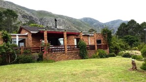 A brick house with a porch, surrounded by a grassy yard and mountains in the background. - Lagoon Edge (Hermanus)