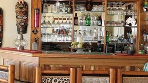 Zona de bar con armarios de madera, estantes con cristalería y una máscara decorativa.