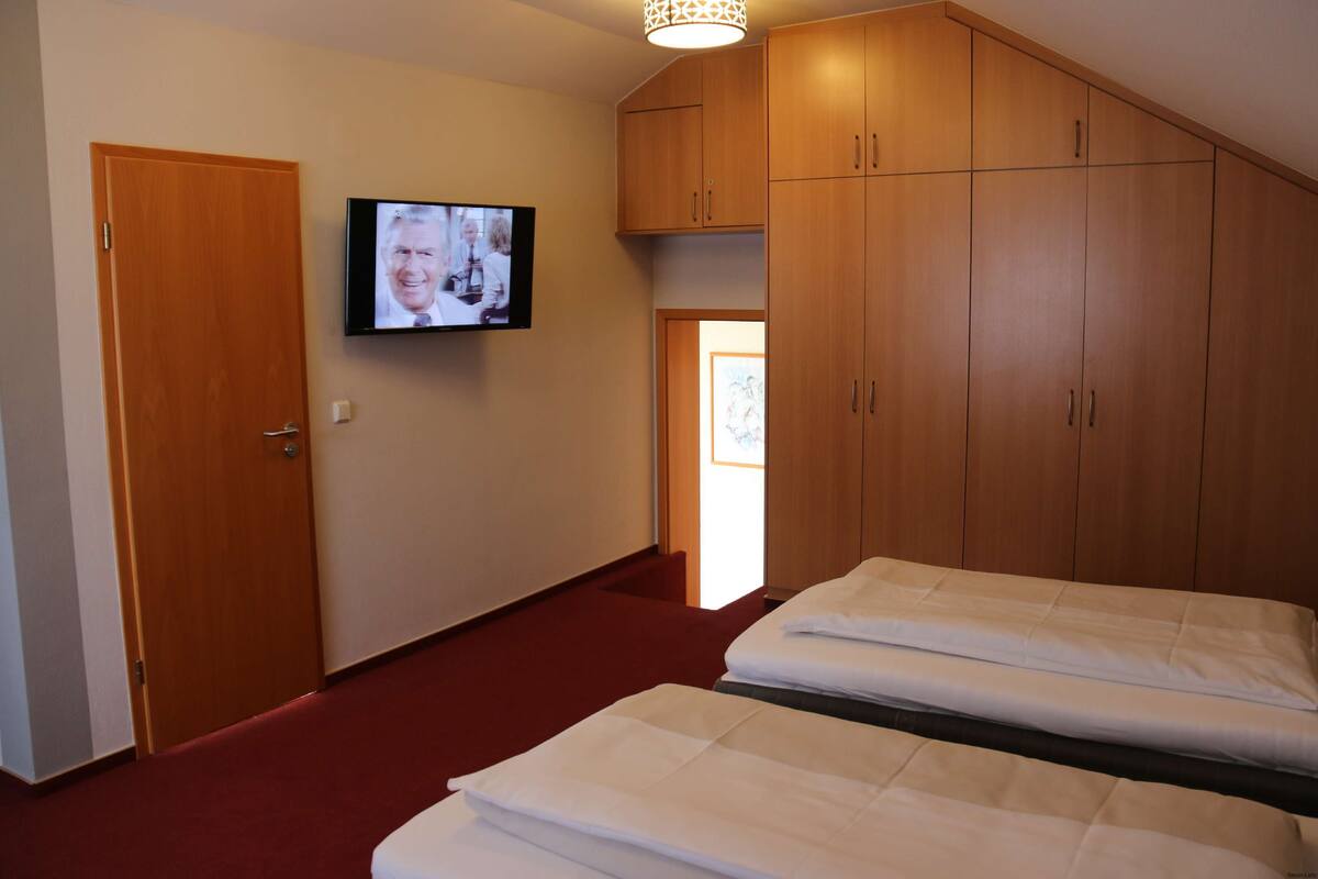 Une chambre d’hôtel avec deux lits, des armoires en bois, un téléviseur fixé au mur et un sol recouvert d’une moquette rouge.