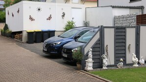 Una zona residencial con un coche azul estacionado, un edificio blanco con techo rojo y una área amurallada con estatuas decorativas.