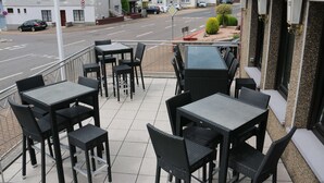 Un patio al aire libre con varias mesas y sillas, con vista a una calle con coches estacionados y edificios residenciales.