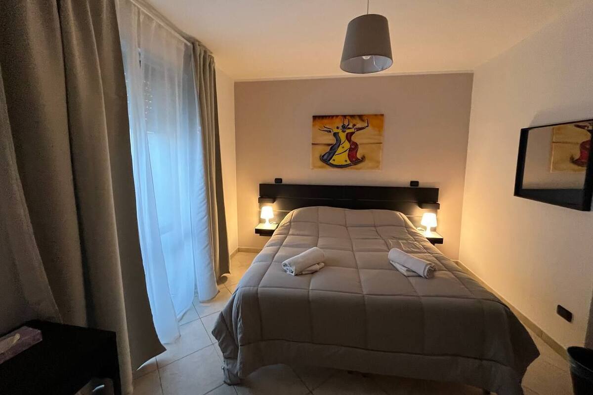Et hotelværelse med en seng, to sengeborde med lamper, et maleri på væggen og et vindue med gardiner.