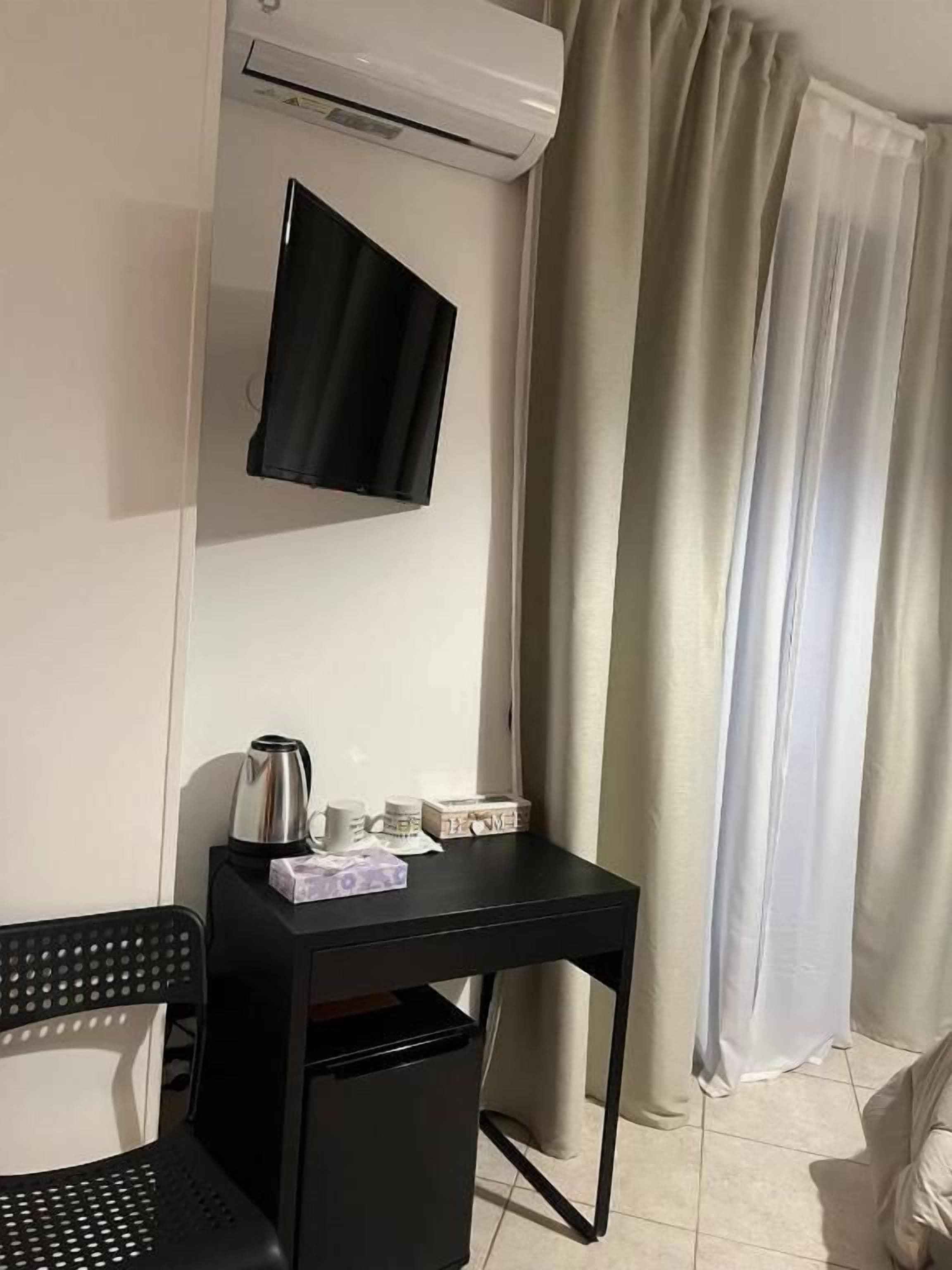 Une pièce avec un bureau noir, une chaise noire, un téléviseur fixé au mur, une petite table avec une bouilloire et des tasses, et une fenêtre avec des rideaux beiges.