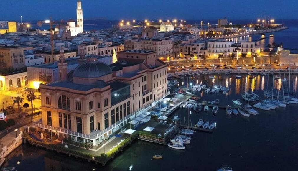A coastal cityscape at night with a prominent building featuring a dome and a waterfront marina.