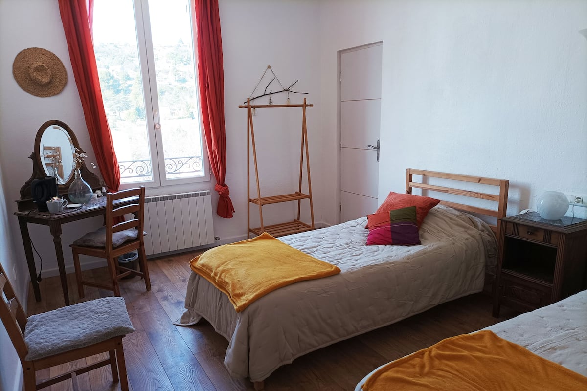 A bedroom with a bed, a wooden chair, a desk with a mirror, a window with curtains, and a radiator.