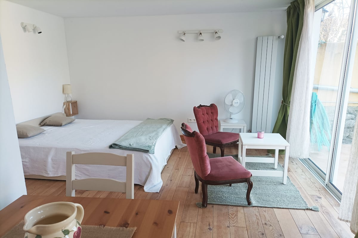 A bedroom with a bed, two chairs, a small table, a fan, and a window with curtains.