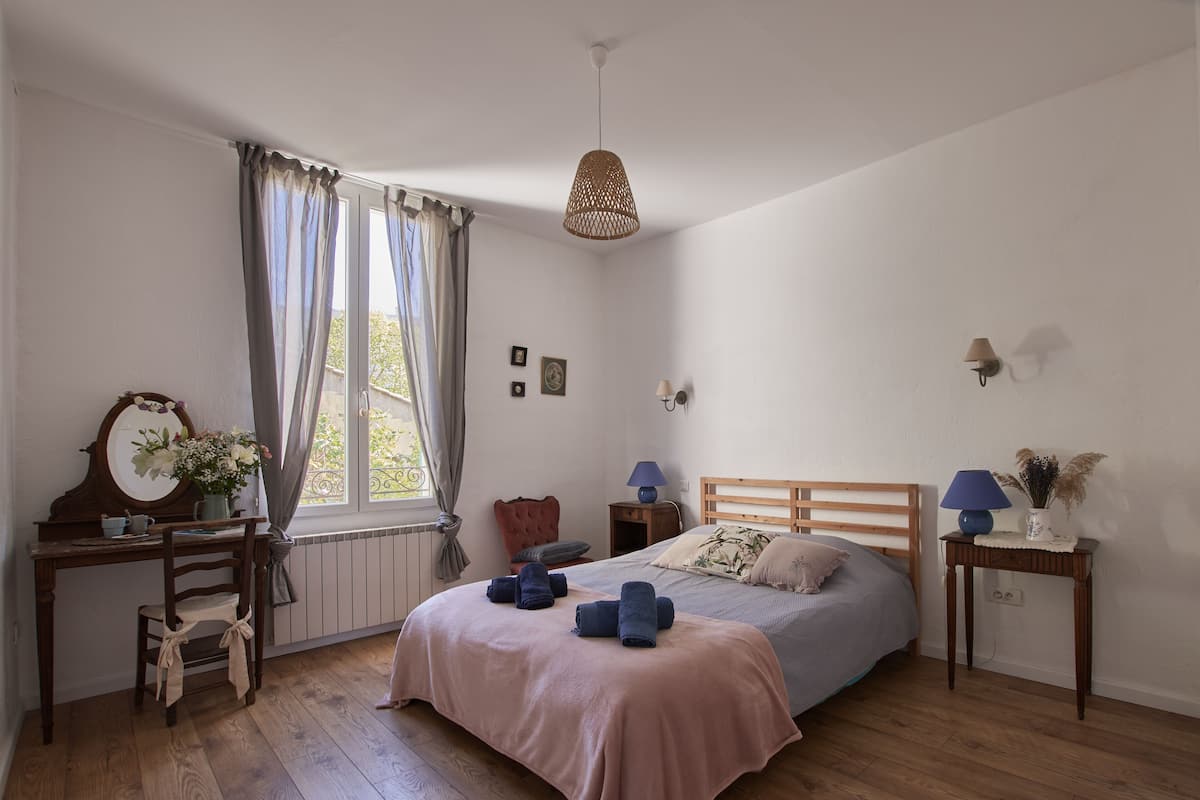 A bedroom with a wooden bed, a desk with a chair, a window with curtains, a bedside table with a lamp, and a wall-mounted light fixture.