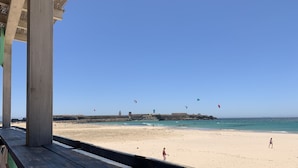 A beach with a wooden deck overlooking the sea, featuring a clear sky and distant rocky formations.