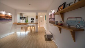 A modern kitchen with a central island, wooden bar counter, and bar stools. There are wooden shelves with decorative items and a small toy van.