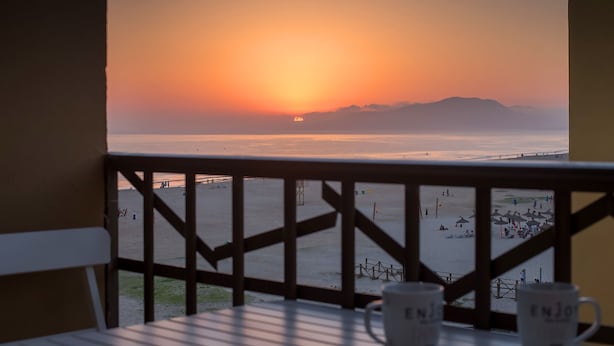 A balcony with a view of a beach at sunset. There are two white mugs on the table.