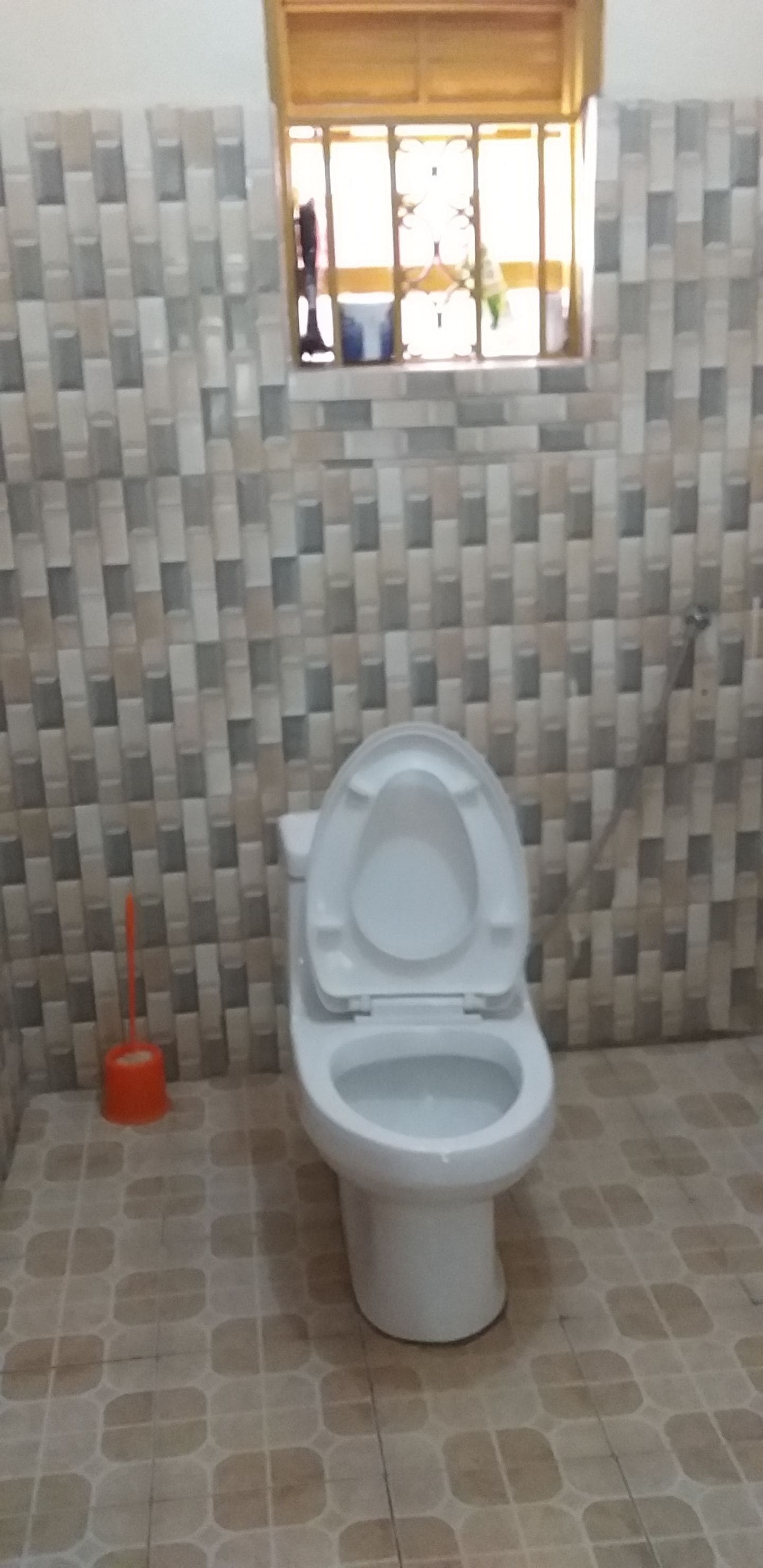 A bathroom with a toilet, a red plunger, and a window with bars.