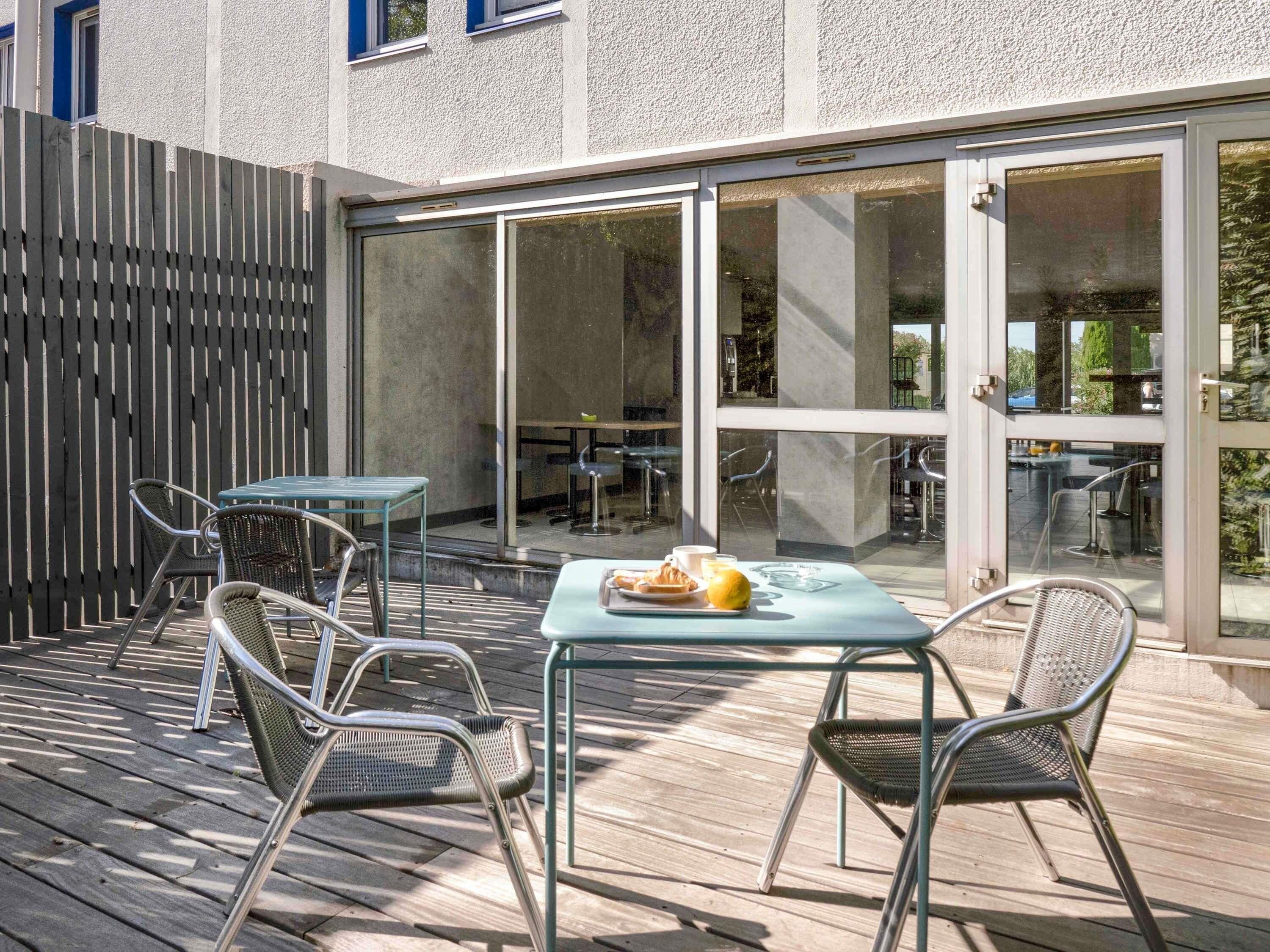 An outdoor dining area with metal chairs and tables, a wooden deck, and a building with large glass doors.