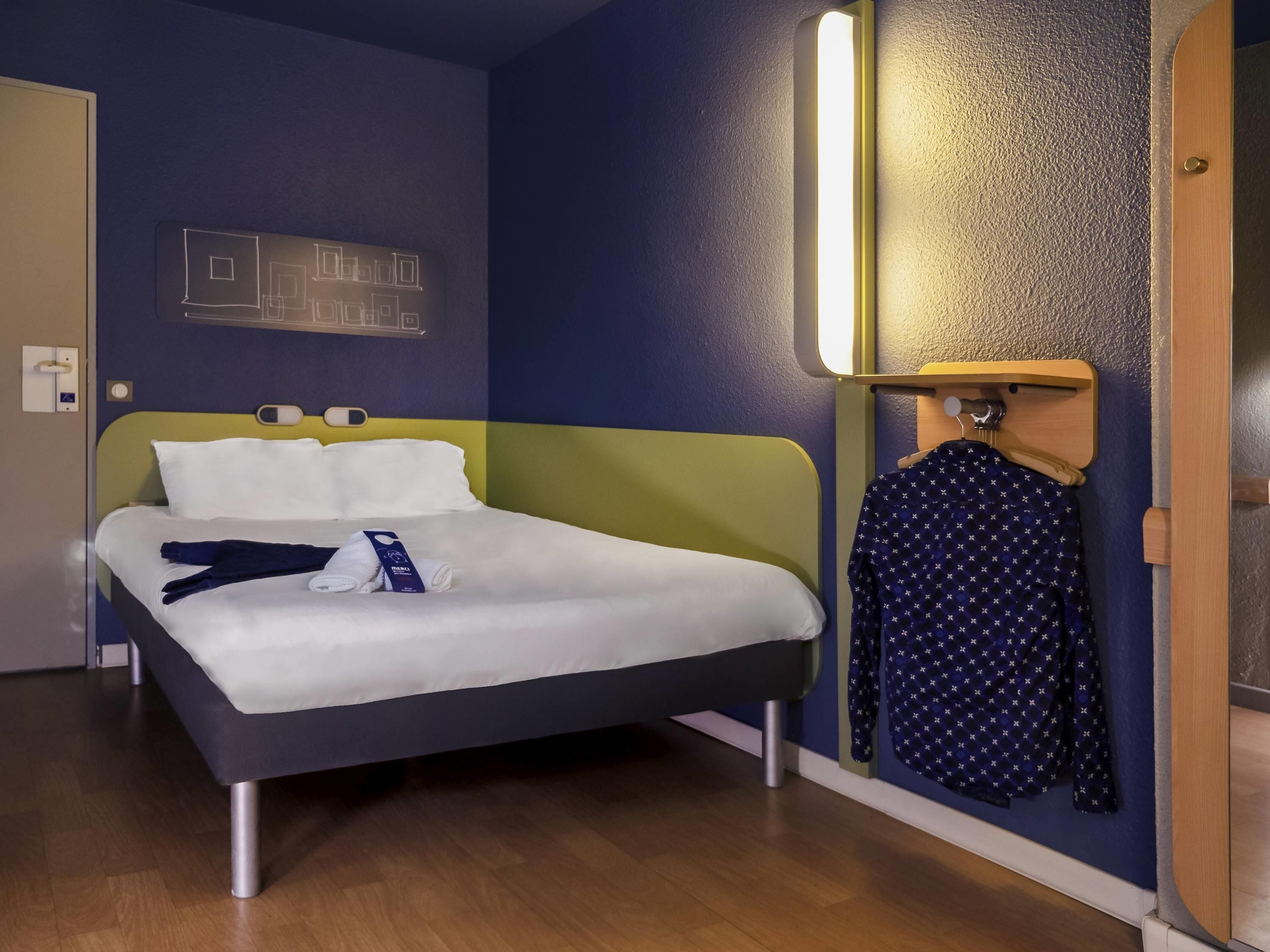 Un lit bien fait, avec du linge de lit blanc, une tête de lit verte et une table de chevet en bois surmontée d’une lampe.