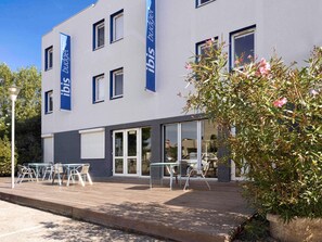 A modern building with a flat roof, large windows, and a covered outdoor seating area with tables and chairs. - Ibis Budget Arles Sud Fourchon (Arles)