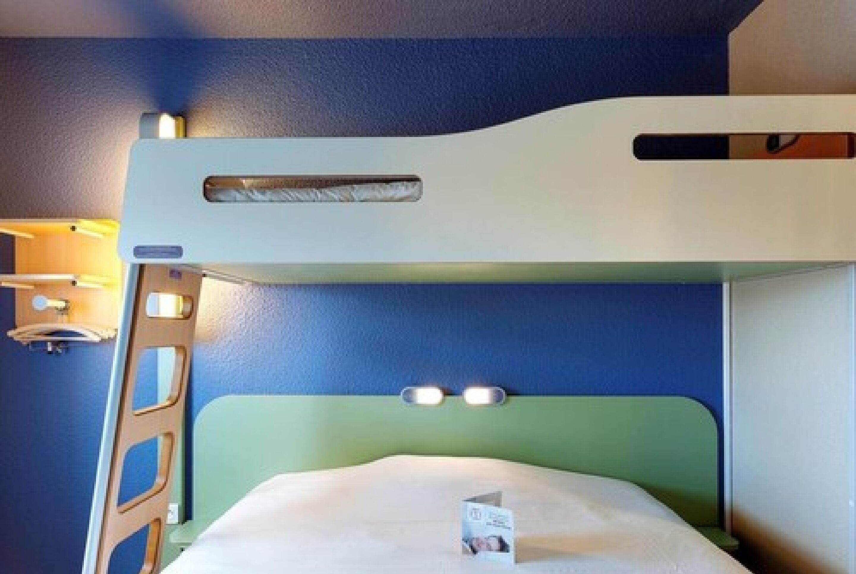 Una cama litera con cabecera verde y colchón blanco. Hay una mesita con una lámpara y una tarjeta encima.
