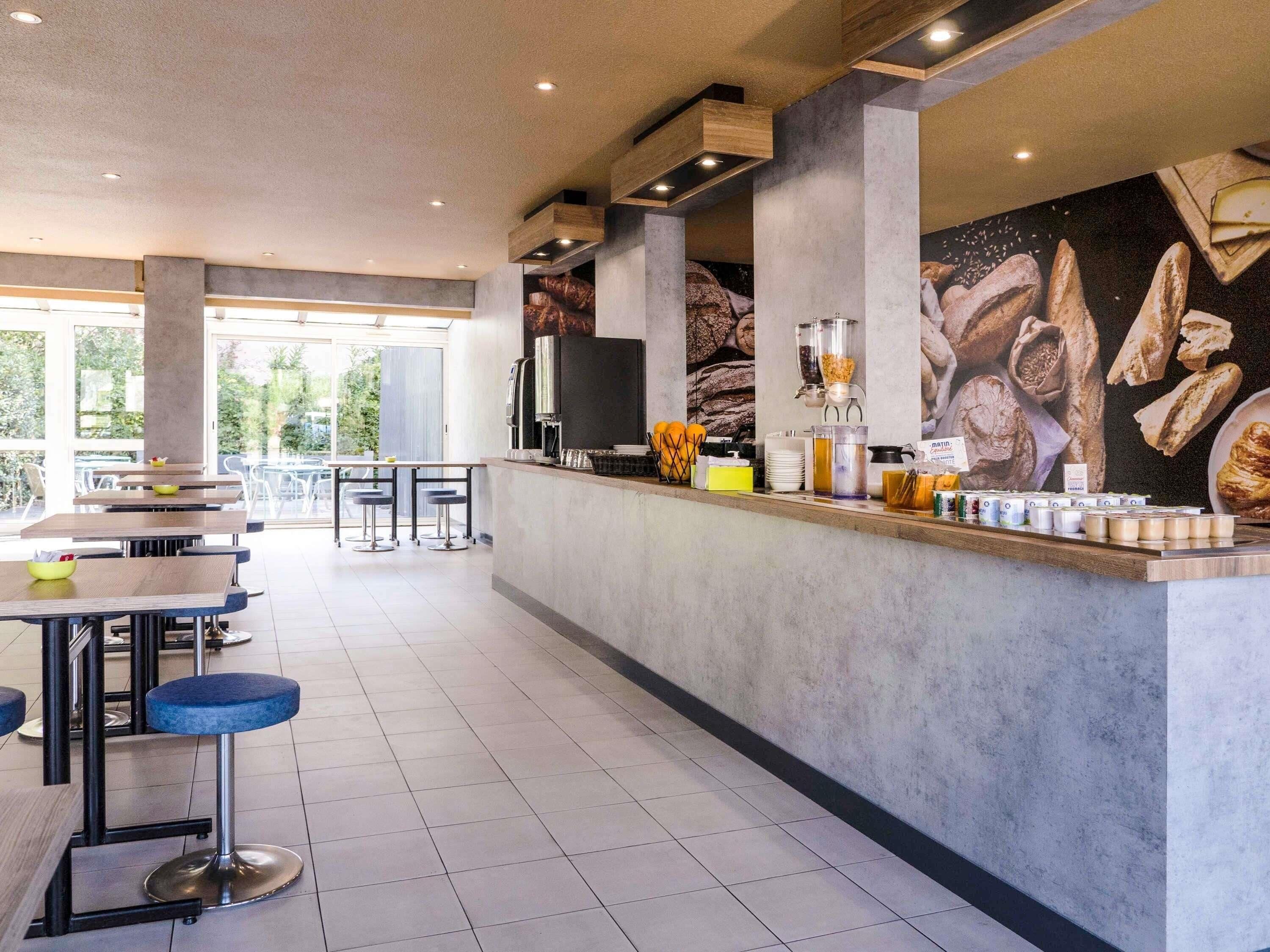Interior moderno de una cafetería con un mostrador central, zona de estar y un amplio ventanal que proporciona luz natural.