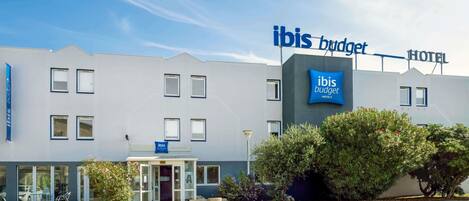 Un moderno edificio de hotel con un llamativo letrero de 'ibis budget', una entrada de vidrio y un estacionamiento.