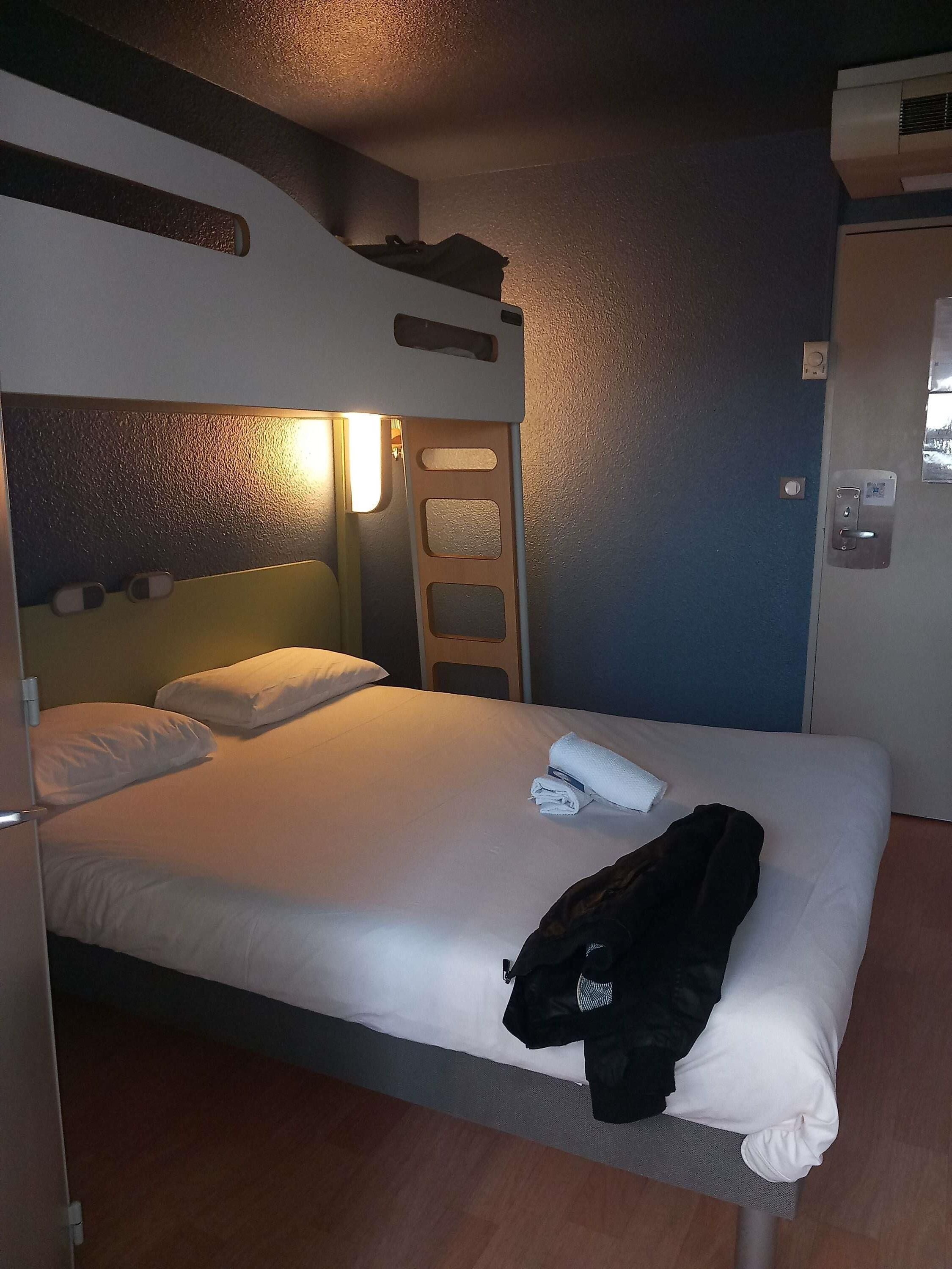 Een eenpersoonsbed met een witte matras en kussens, een opgerolde witte handdoek en een zwart jack op het voeteneinde.