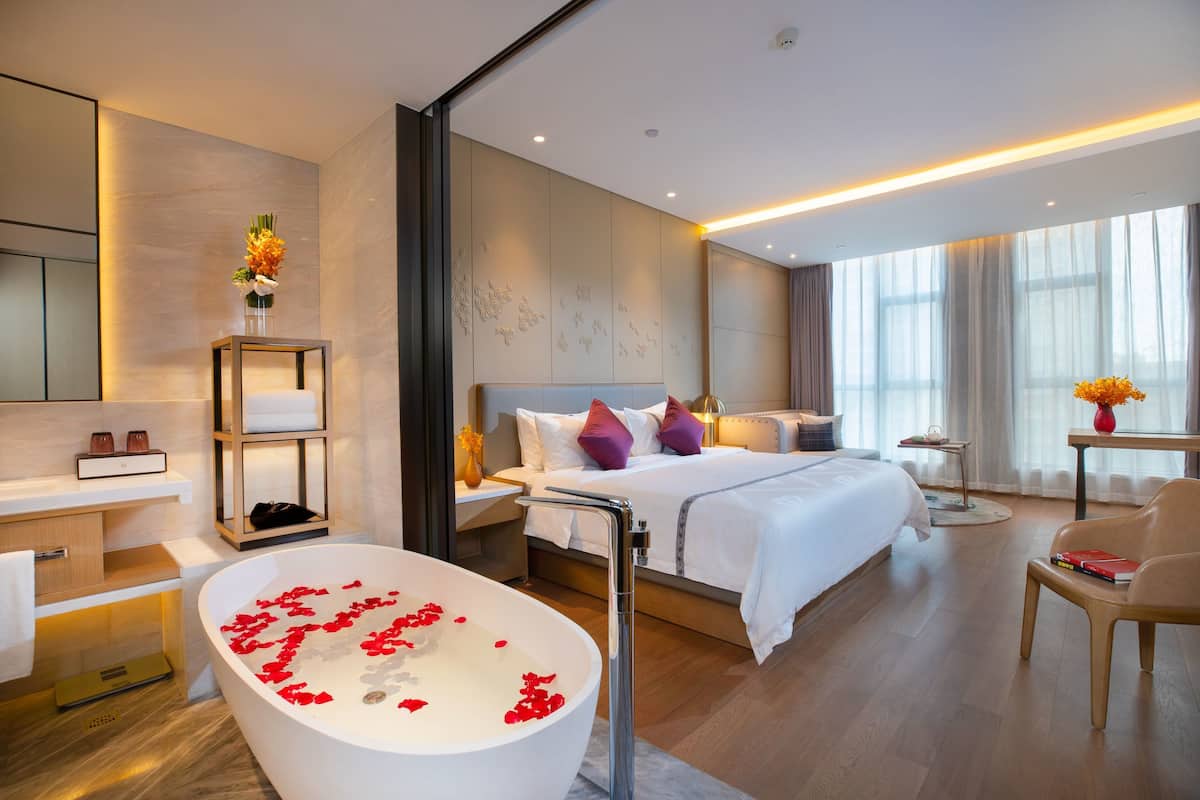 Una habitación de hotel moderna con una cama grande, una bañera con pétalos de rosa, un rincón de descanso y un ventanal amplio.