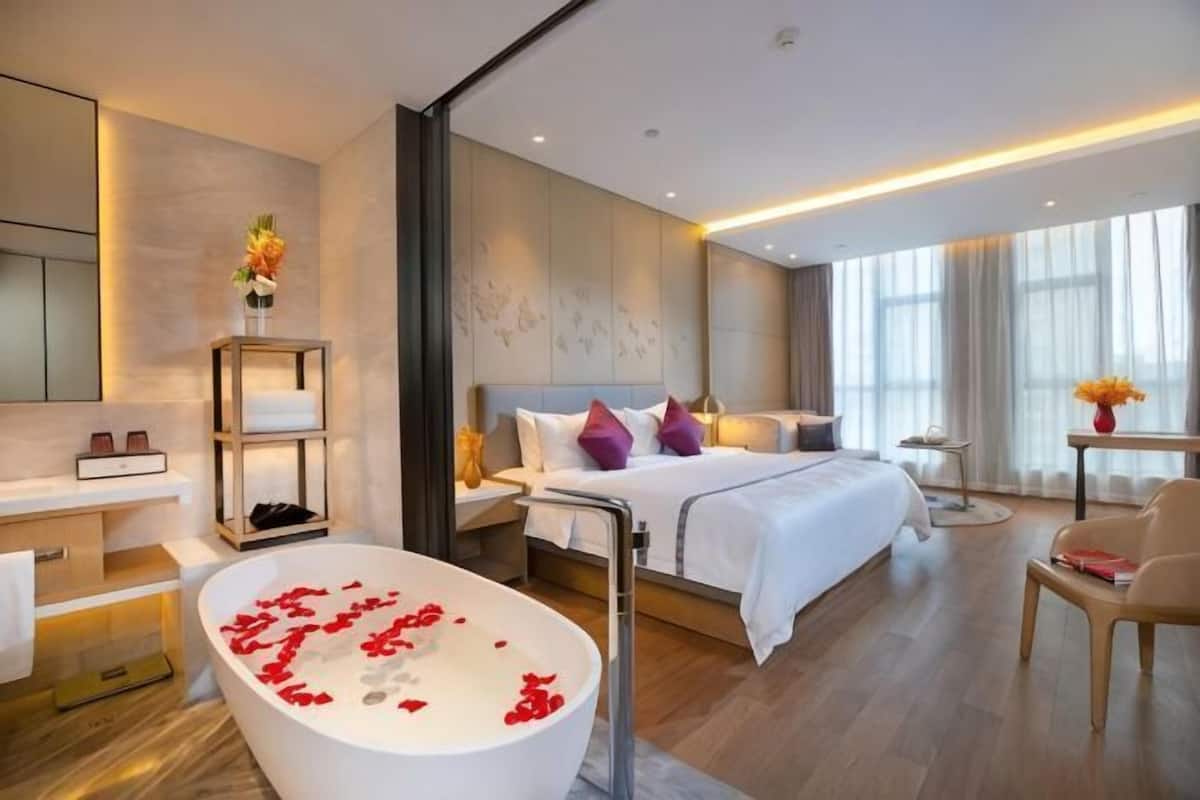 Una habitación de hotel moderna con una cama grande, una bañera con pétalos de rosa, una zona de estar y vistas a la ciudad.