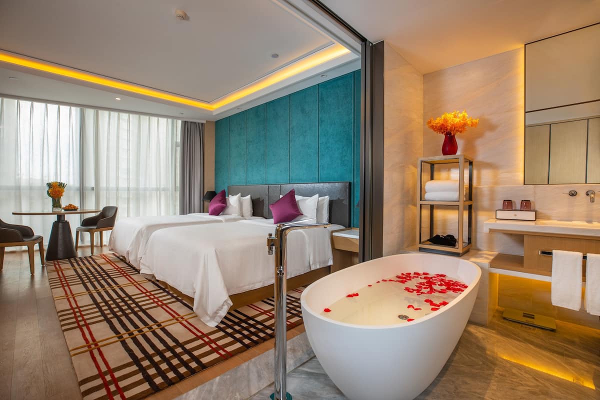 Una habitación de hotel moderna con una cama grande, una zona de estar con sillas, una bañera con pétalos de rosa y un baño con espejo y perchero.