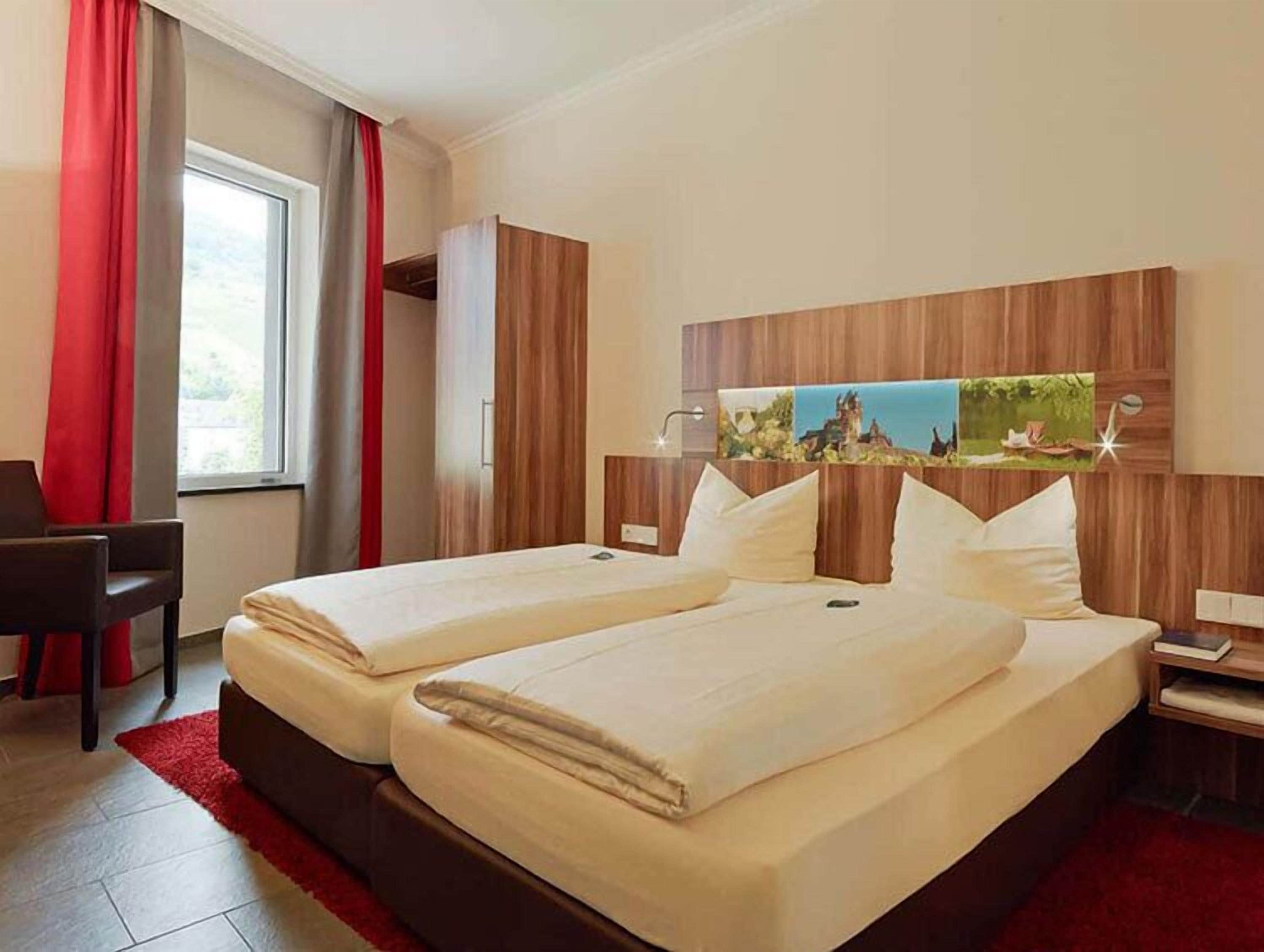 Ein Hotelzimmer mit zwei Betten, einem Schreibtisch und einem großen Fenster mit roten Vorhängen.