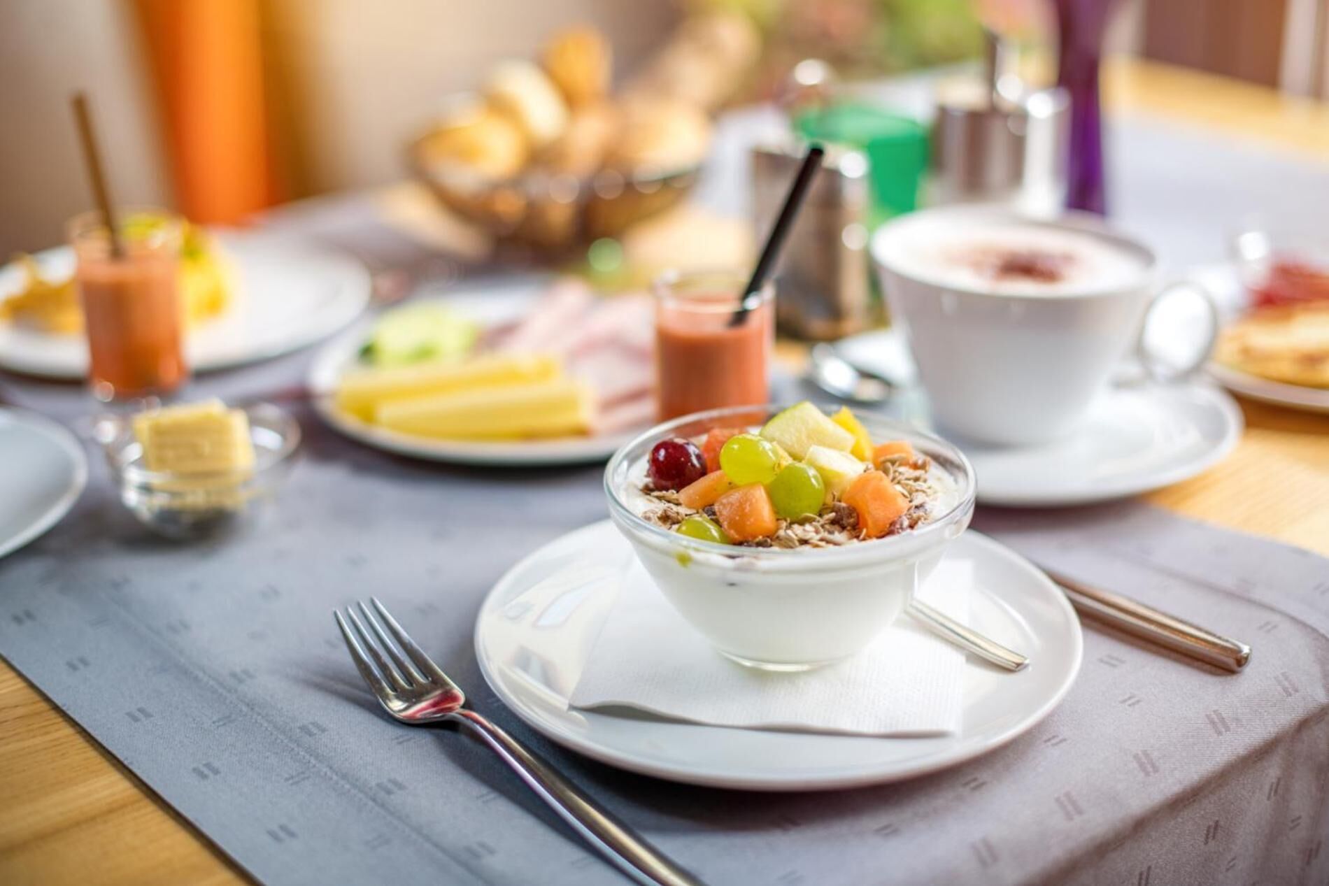A breakfast setting with a bowl of yogurt topped with fruits and granola, a cup of coffee, and various pastries on the table.