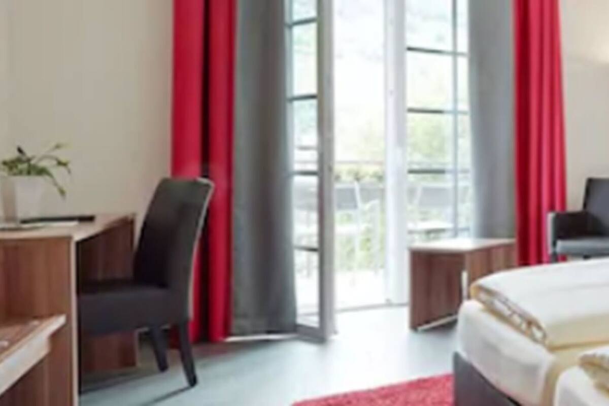 Habitación de hotel con dos camas, un escritorio, una silla y un ventanal con cortinas rojas.