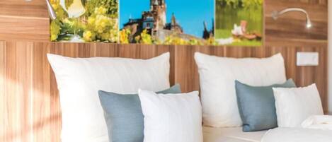 Habitación de hotel con cabecera de madera, cama con ropa de cama blanca y almohadas azules y blancas, y un cuadro en la pared que muestra un castillo y follaje verde.