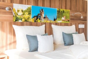 Quarto de hotel com cabeceira de madeira, cama com roupa de cama branca e almofadas azuis e brancas, e uma imagem pendurada na parede retratando um castelo e vegetação.