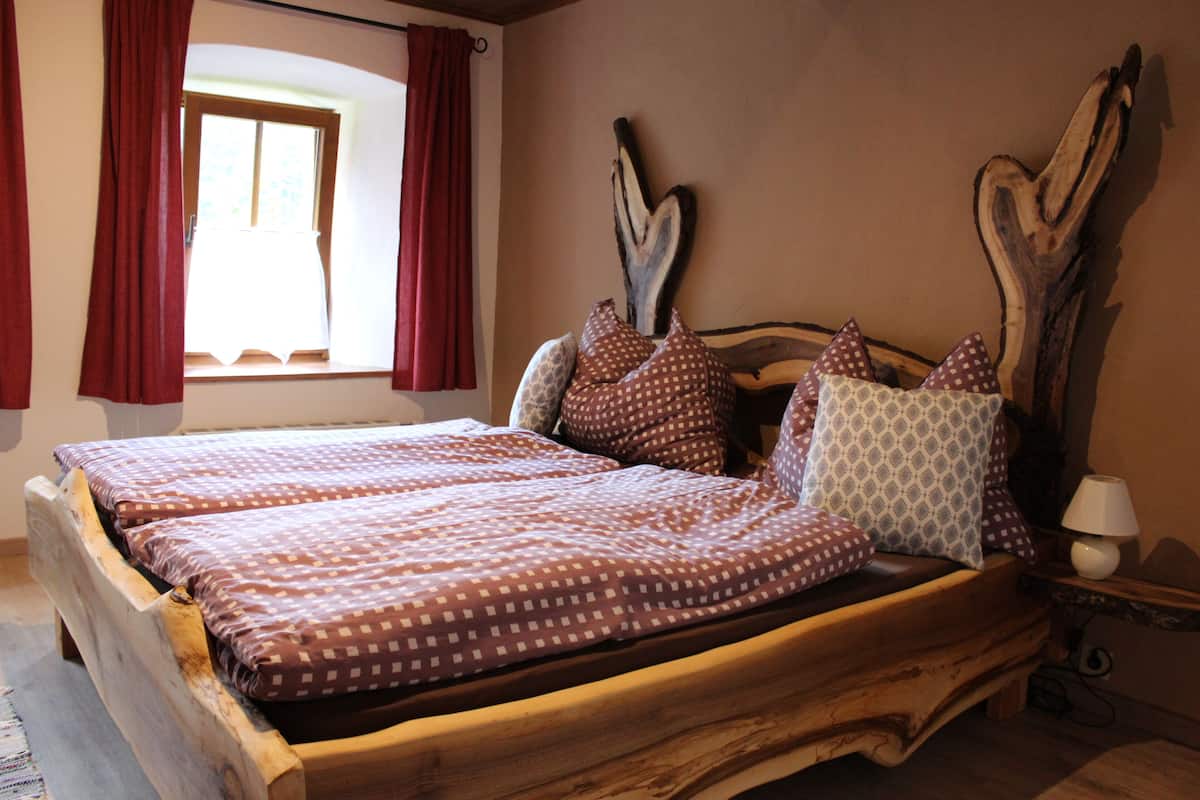 Ein hölzernes Bett mit gemusterten Textilien, ein Nachttisch mit einer Lampe und ein Fenster mit roten Vorhängen.