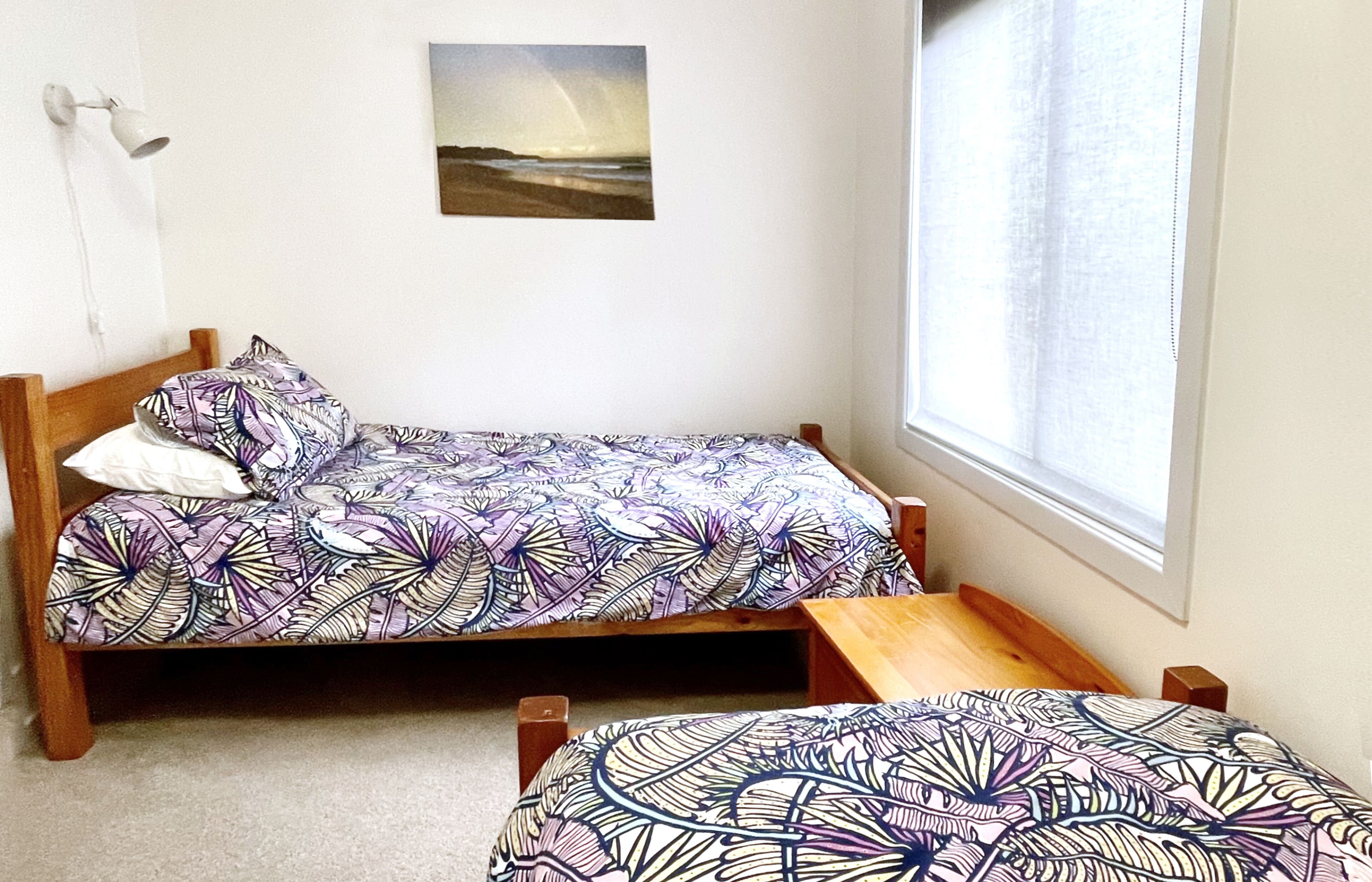 A single bed with a patterned bedspread and pillows, a small wooden bench, a wall-mounted lamp, and a framed picture of a beach scene.