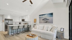 Ett modernt kök med vita köksskåp, en köksö med barstolar, en vit soffa och en tavla med strandmotiv på väggen.