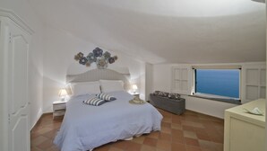 A bedroom with a bed, a nightstand, a lamp, a decorative wall piece, a window with a view, and a small table. - Casa Malu' (Praiano)